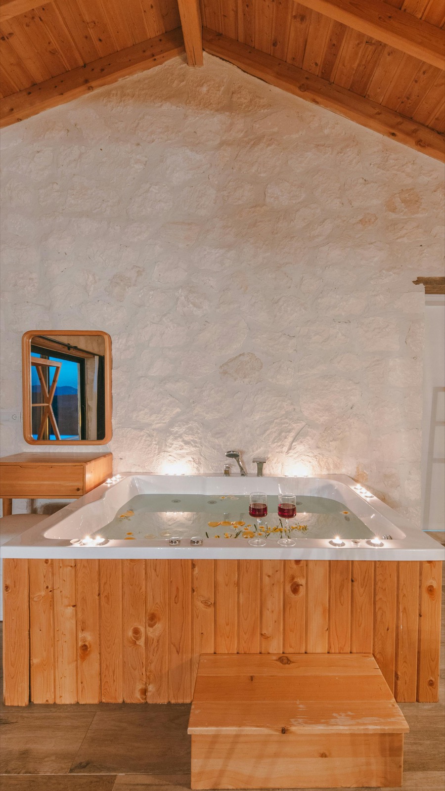 A rectangular spa in a wood-roofed cabin with white stone walls — two glasses of red wine resting on the tub edge, candles lit around the water.