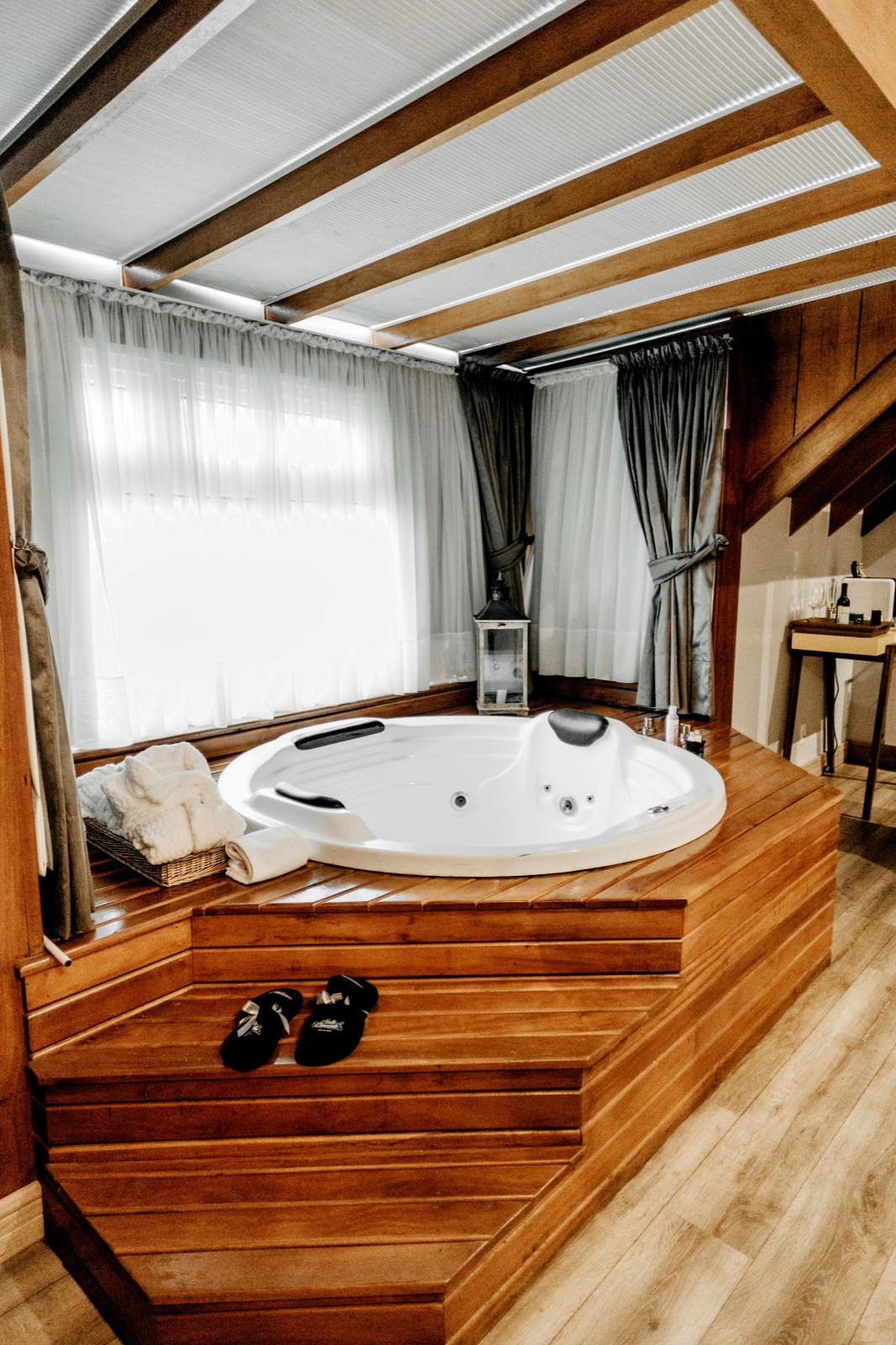 A round whirlpool spa set into a wooden deck inside a chalet — beamed ceiling, sheer curtained windows, slippers waiting on the steps.